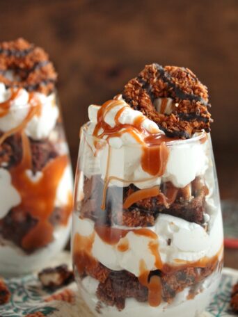 Layers of whipped cream, brownies, caramel, and Samoa cookies in stemless wine glass