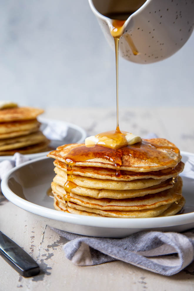 Two stacks of banana sourdough pancakes on a white background with maple syrup being poured on one stack