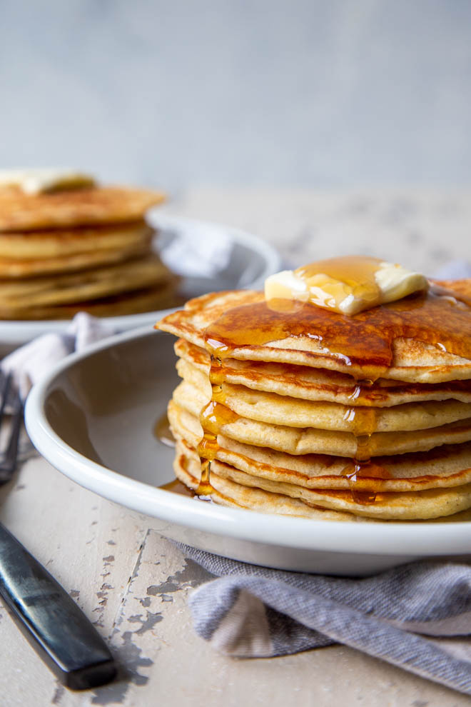 Two stacks of banana sourdough pancakes on a white background, a close up shot of one stack with maple syrup falling over the edges
