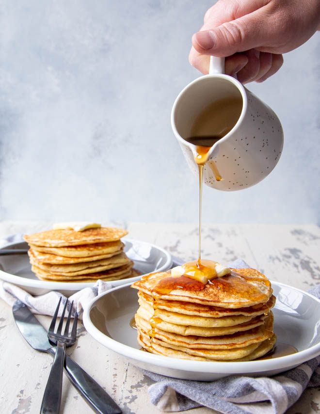 Two stacks of banana sourdough pancakes on a white background with black matte utensils and syrup being poured over one stack