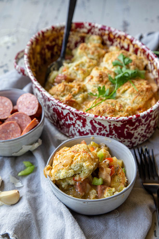 Full on comfort food with andouille sausage and chicken pot pie, topped with cheesy herbed biscuits in baking dish and one serving in a gray bowl