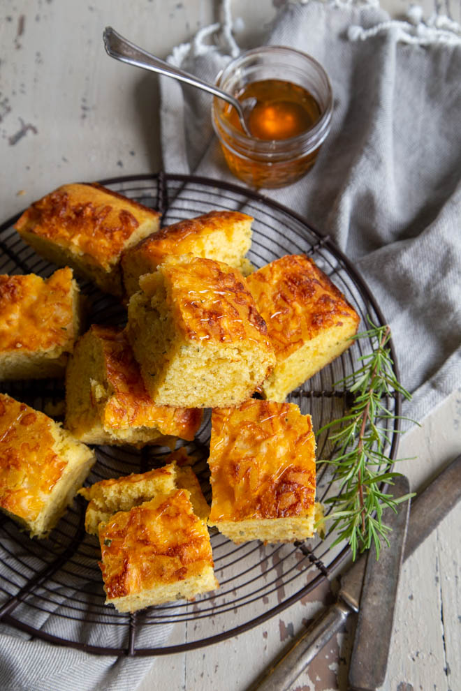 square slices of cornbread on a wire rack with a sprig of rosemary and jar of honey