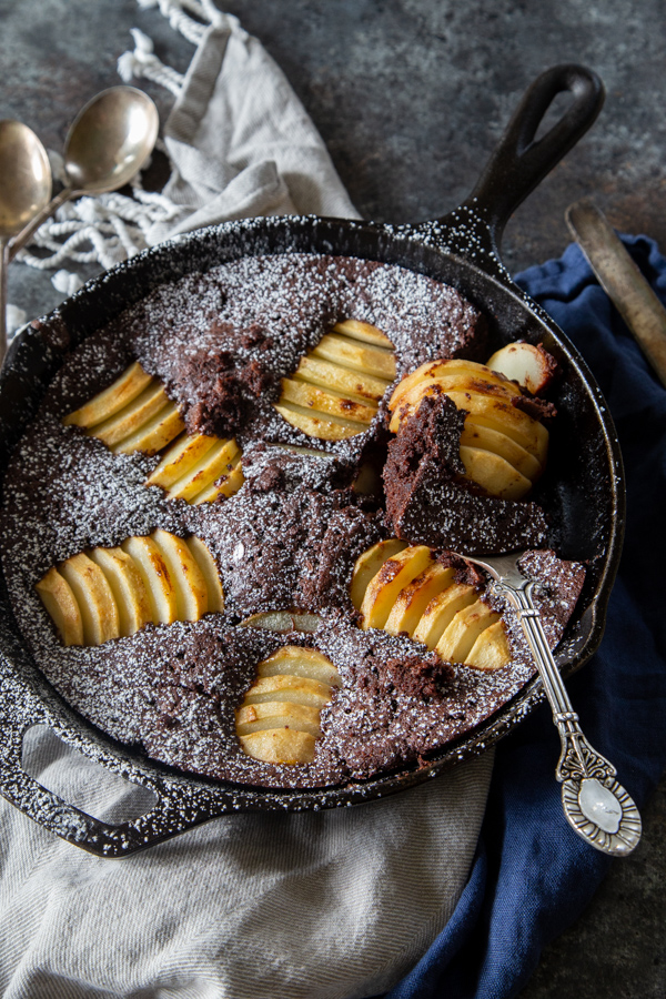 Chocolate chai and pear skillet cake in a cast iron skillet on a gray and blue napkin shot overhead with a slice removed.