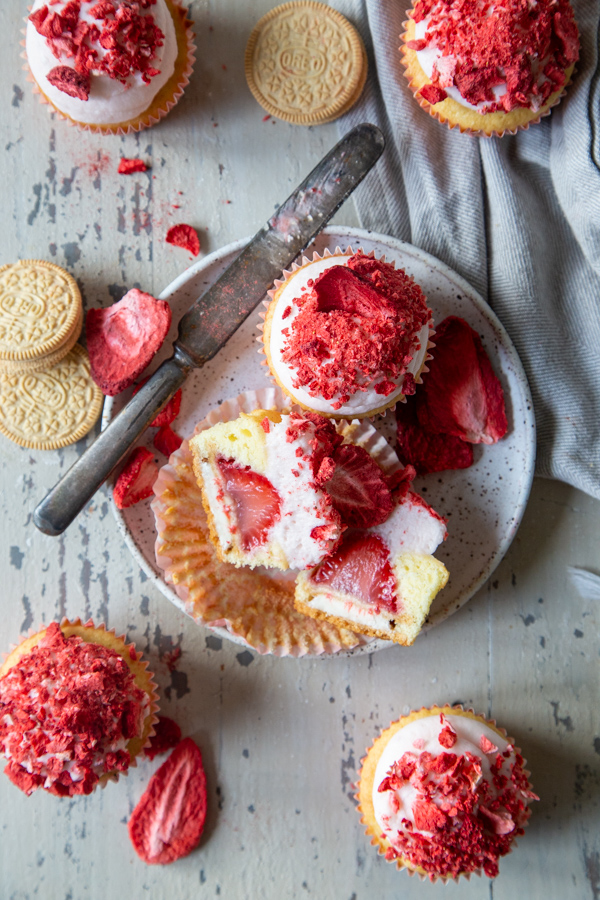 overhead photo of strawberry shortcake cupcakes, with some whole and one cut in half on a white speckled plate