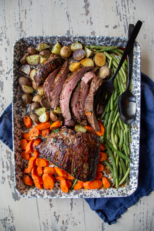 sheet pan steak and veggies dinner on a gray and white sheet pan on a white distressed table.