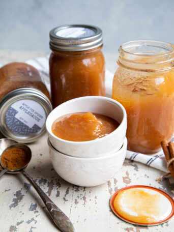 Three glass mason jars sitting behind two nested white ceramic bowls filled with applesauce and spoon holding ground cinnamon