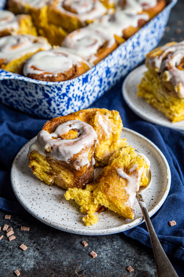 Two pumpkin cinnamon rolls on plates with baking dish of rolls in background