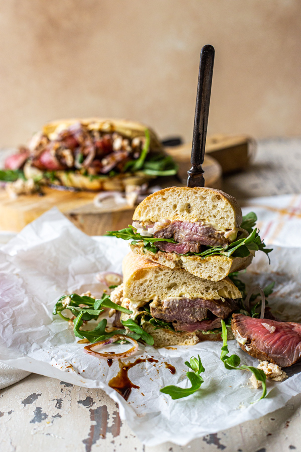 Sirloin steak sandwich sliced in half and stacked, with a knife supporting it. Sliced of steak and arugula around it.