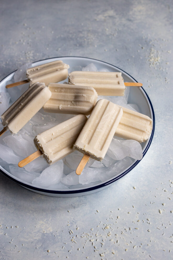 Vegan coconut paletas on a bowl of ice and white background with coconut shreds all around.