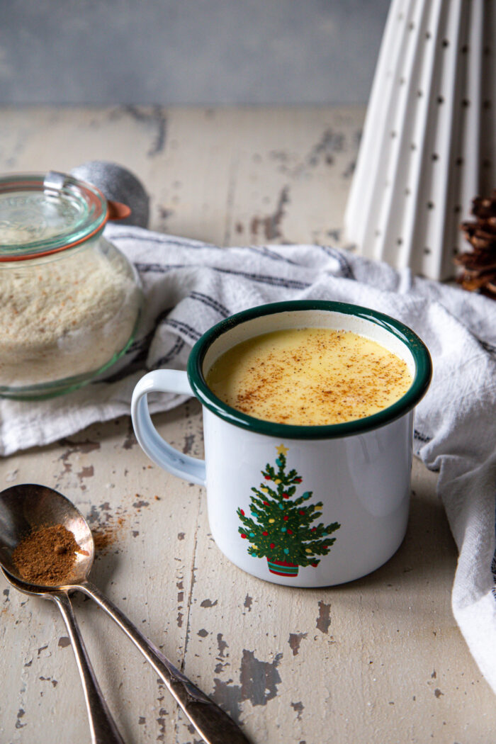 eggnog white chocolate hot cocoa in a white enameled mug with a christmas tree on it with nutmeg grated on top of the cocoa