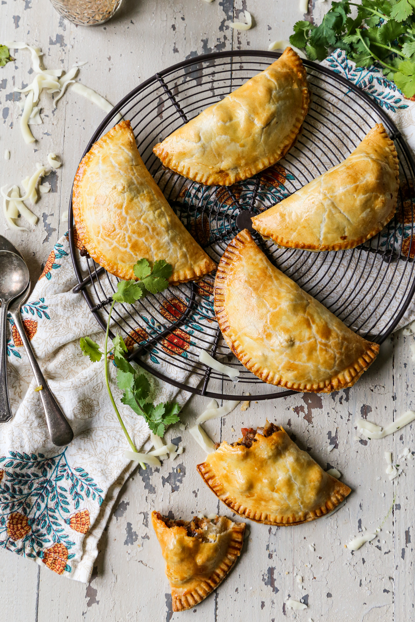 cast iron beef empanadas on a cooling rack with cilantro and cheese sprinkled around on a white background