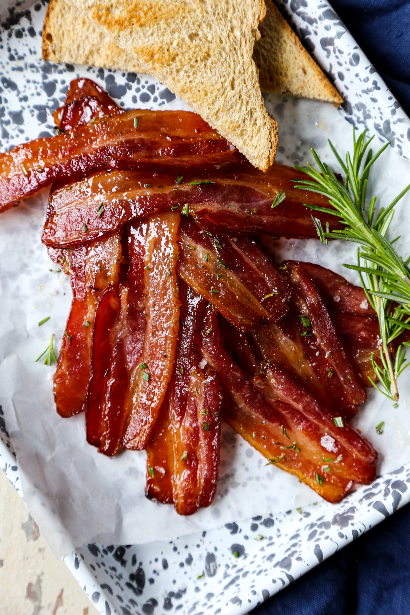 Maple rosemary bacon cooked in the oven on a parchment lined baking sheet with rosemary sprigs and toast