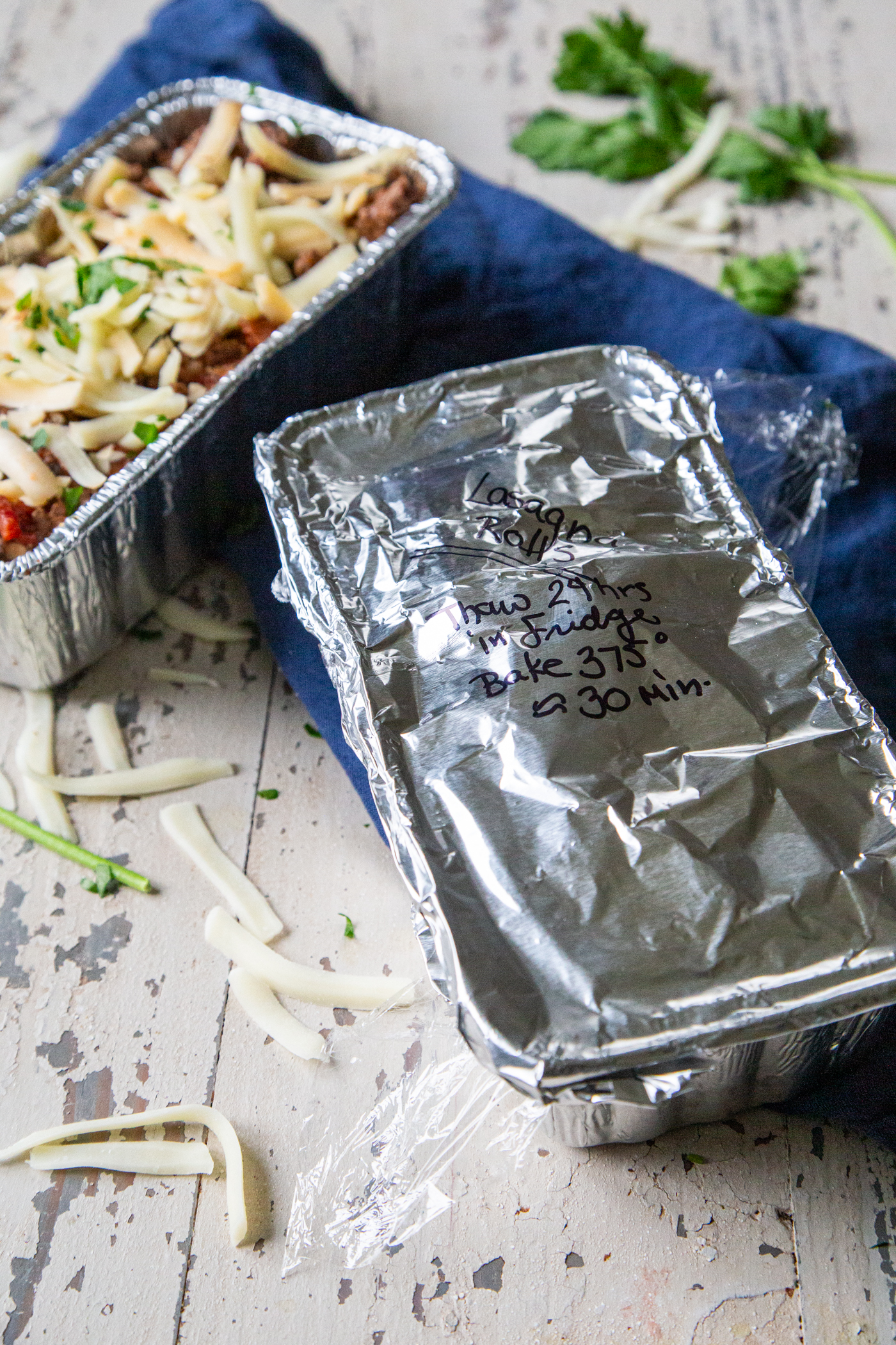 Overhead image of a lasagna rolls filled aluminum loaf pan topped with cheese and one covered in foil with baking directions written on top