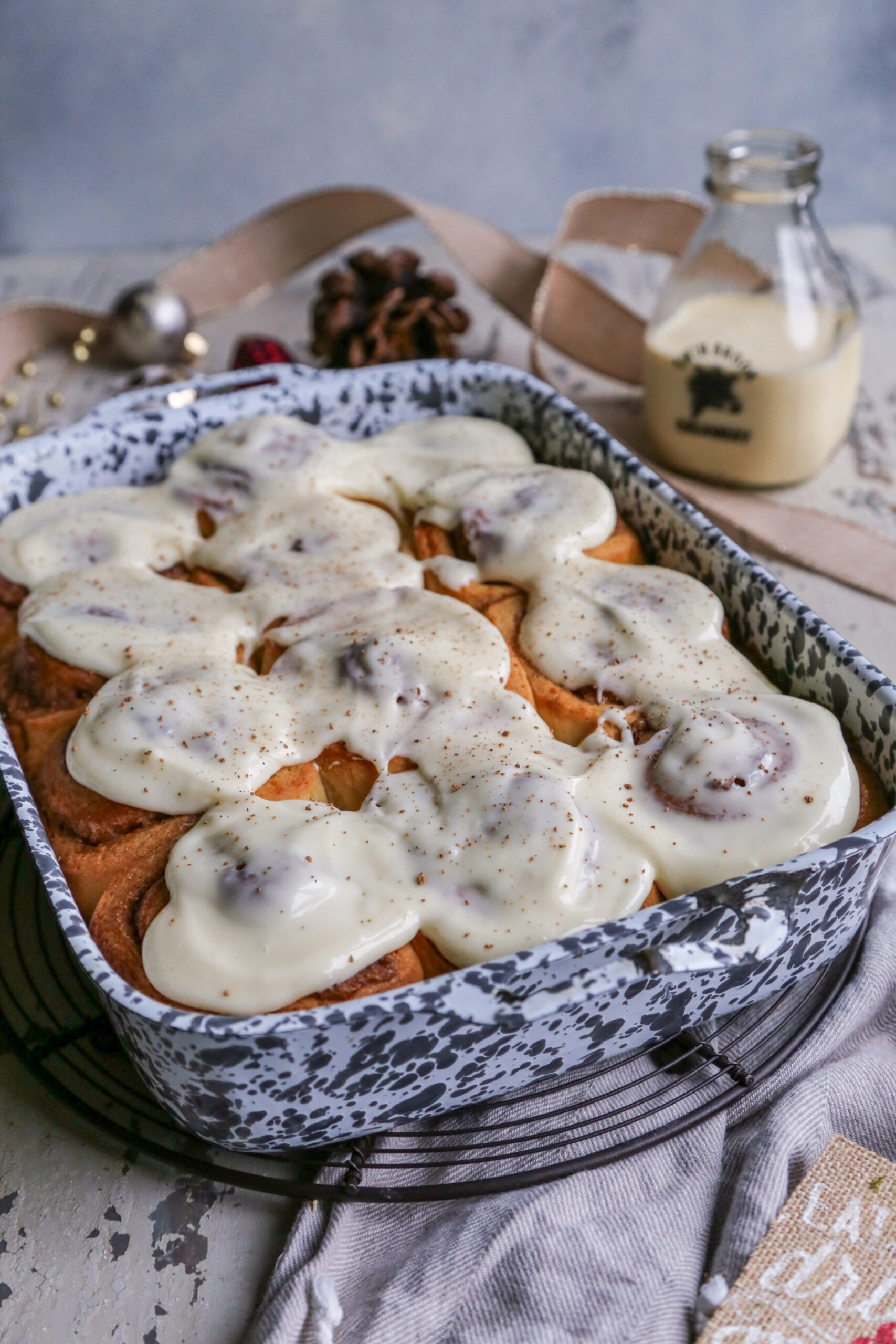 Photo of eggnog cinnamon rolls in a gray and white speckled baking dish, on a rustic white background with Christmas ornaments around it. 