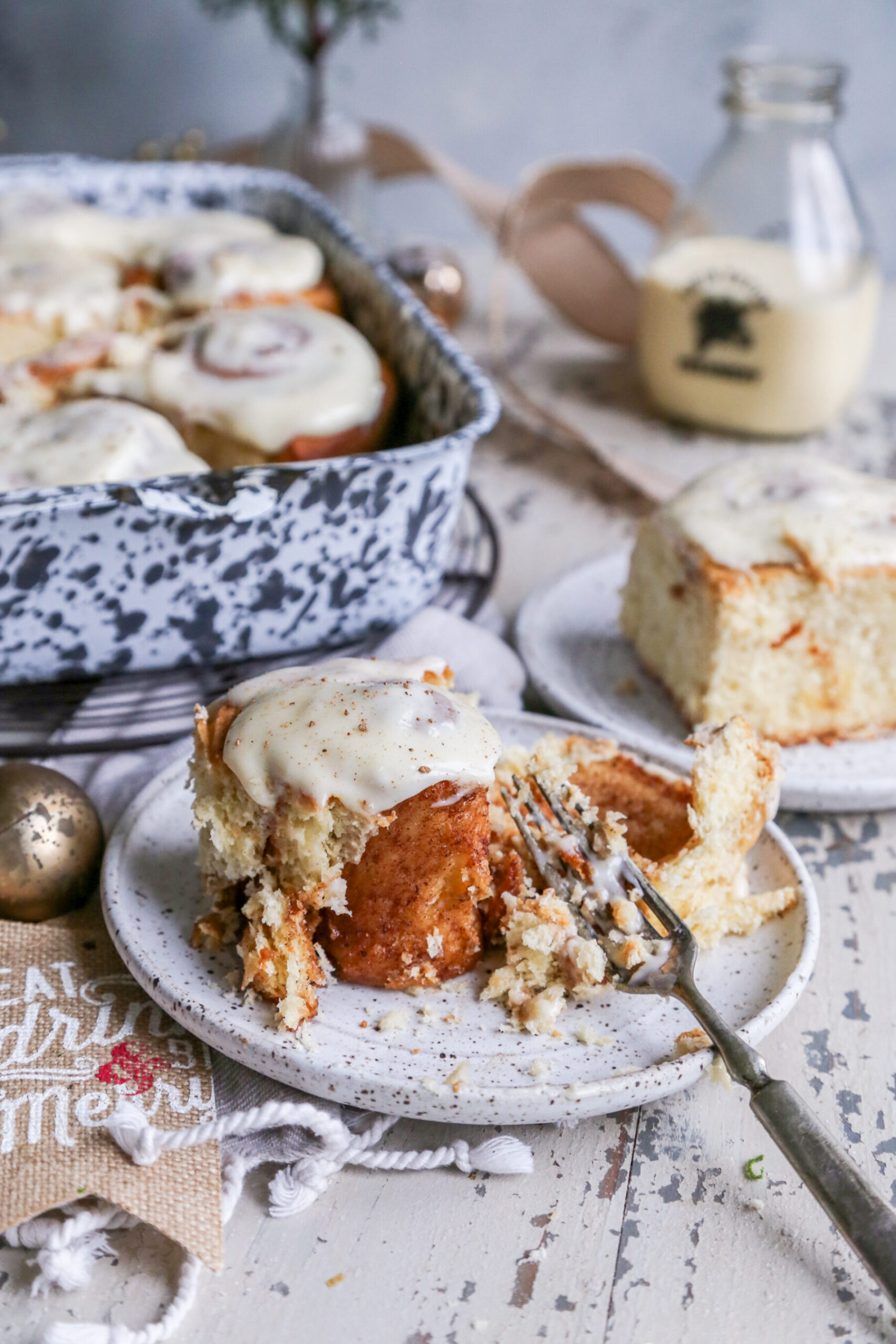 Photo of an eggnog cinnamon rolls on a small plate and in a gray and white speckled baking dish, on a rustic white background with Christmas ornaments around it. 