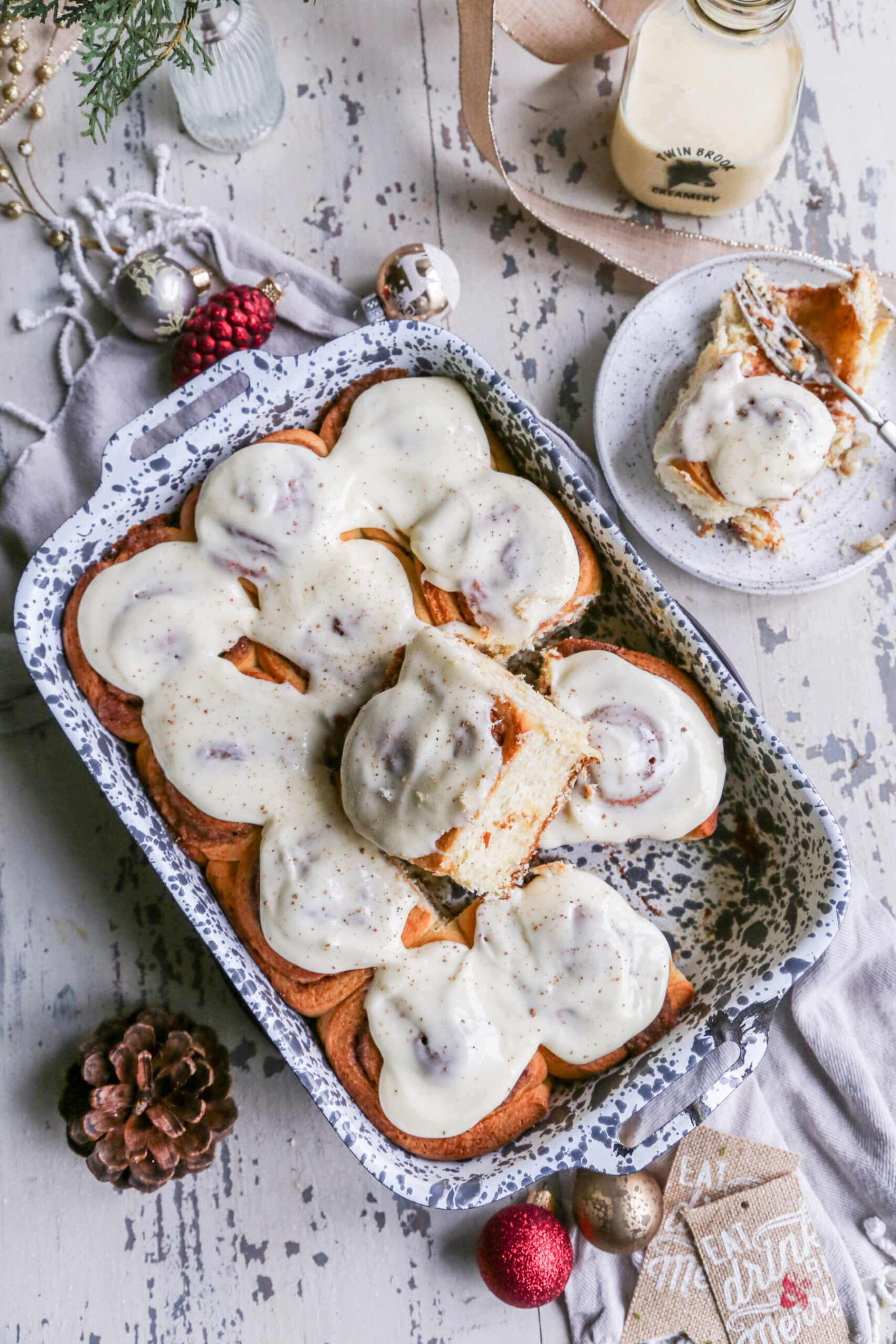 Overhead photo of eggnog cinnamon rolls in a gray and white speckled baking dish with one sticking out, on a rustic white background with Christmas ornaments around it. 