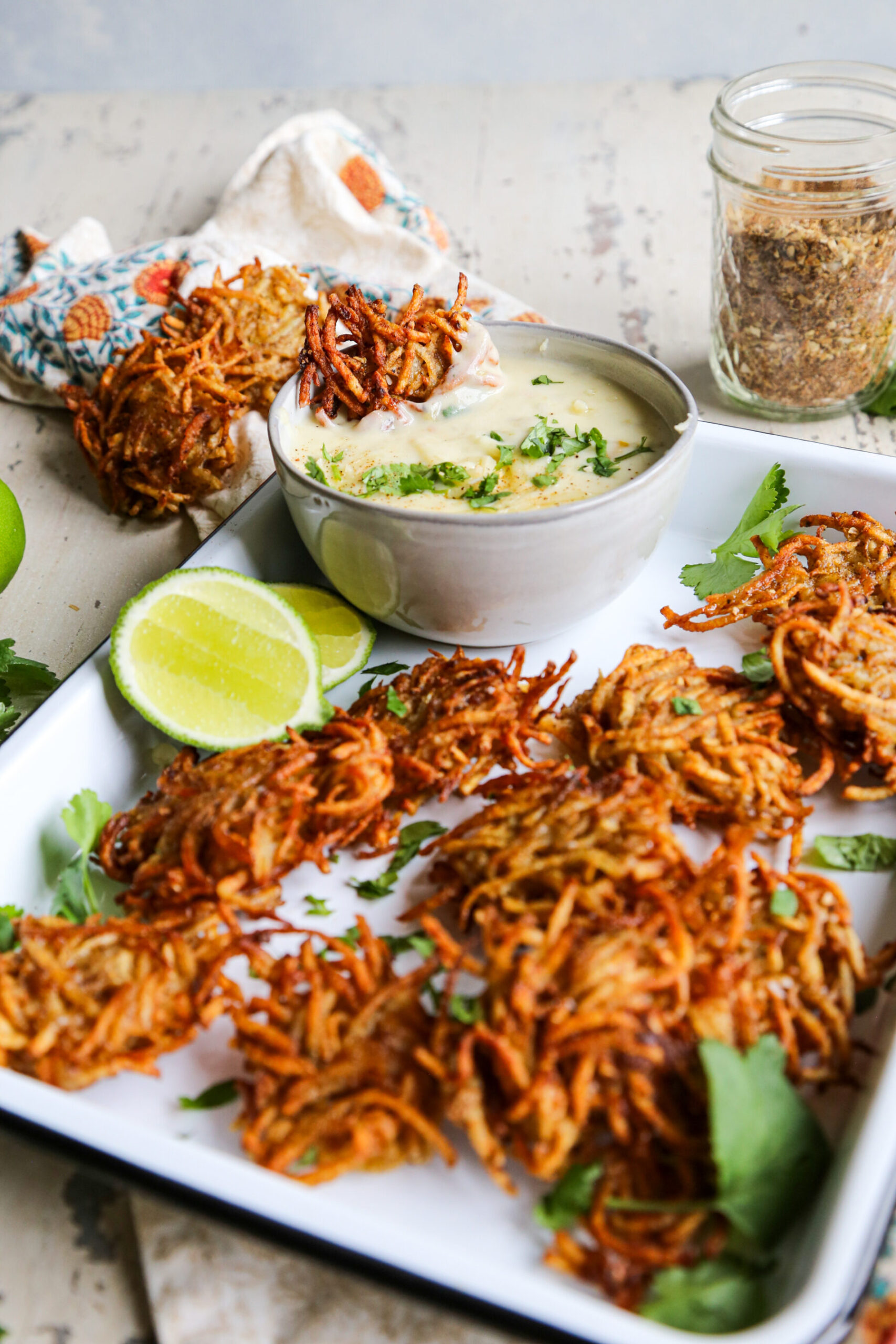 Angled shot of taco spiced potato fritters / latkes on a white baking sheet with a bowl of cheese dip and lime