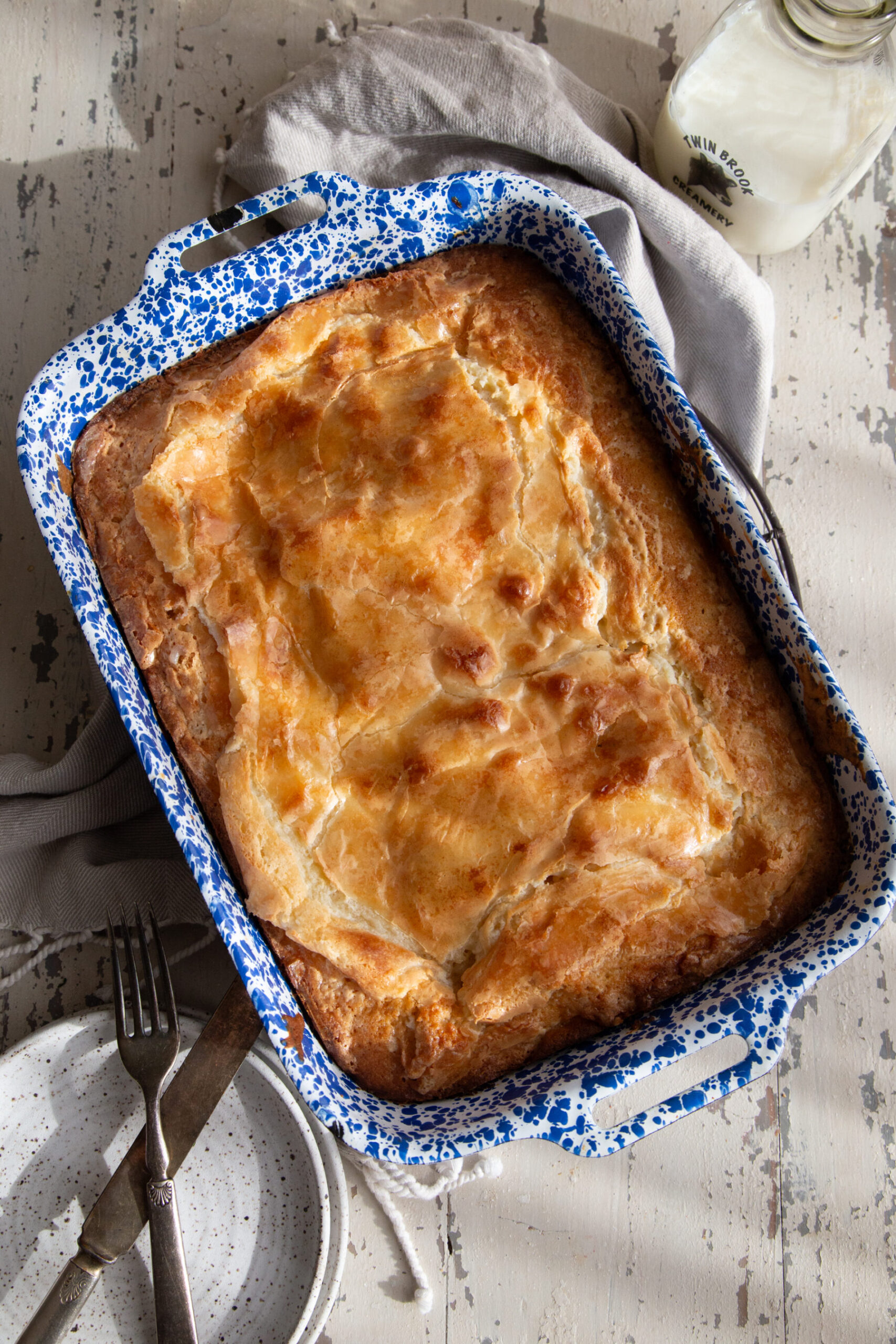 A baked ooey gooey butter cake in a blue and white speckled baking dish, from overhead, flanked by a bottle of milk and a speckled plate with antique knife and fork. 