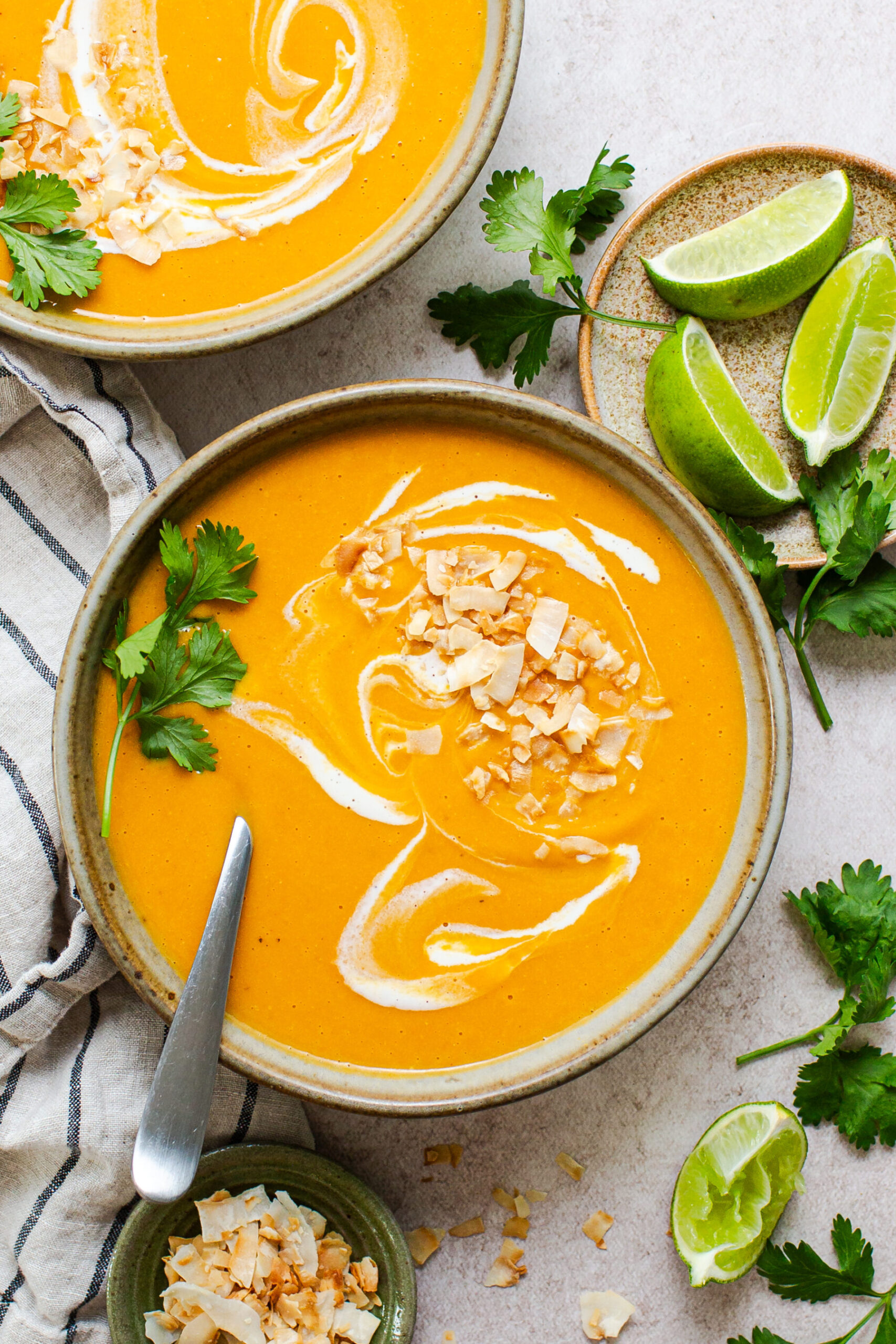 Bowl of butternut squash soup with toasted coconut on top and a swirl of cream to contrast. In an earthenware bowl shot from overhead, surrounded by lime wedges and cilantro leaves.